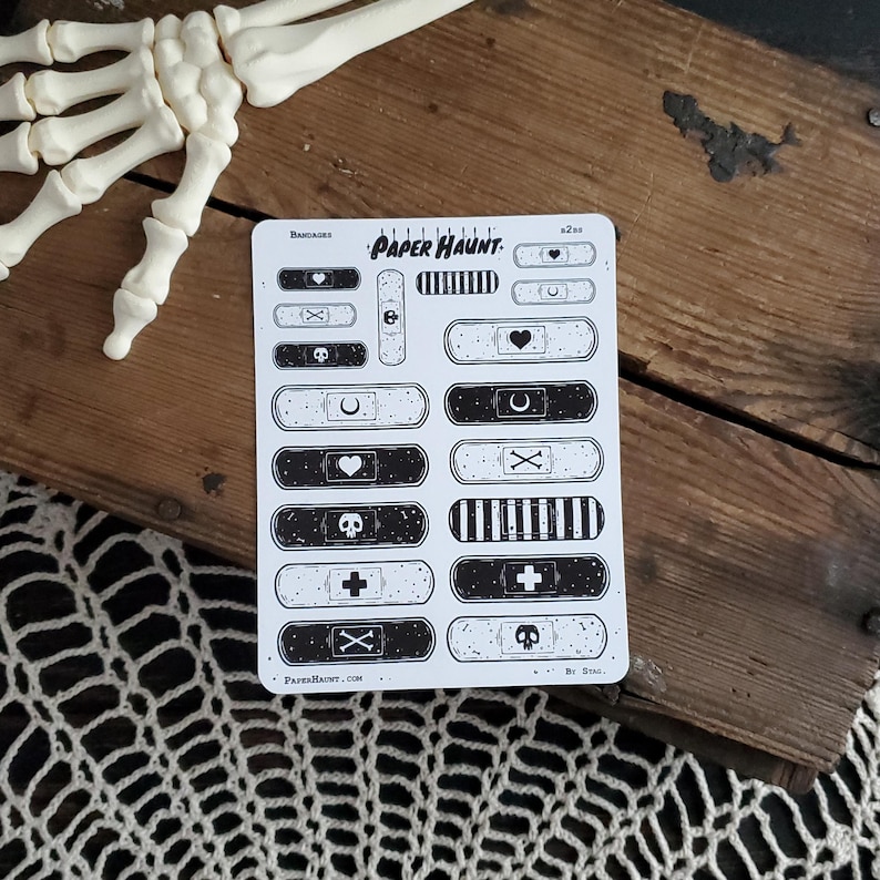 May include: A sheet of black and white illustrated band-aid stickers with various designs, including skulls, crosses, hearts, and crescent moons. The sheet is titled "Paper Haunt" and has the text "Band-Aids" at the top.