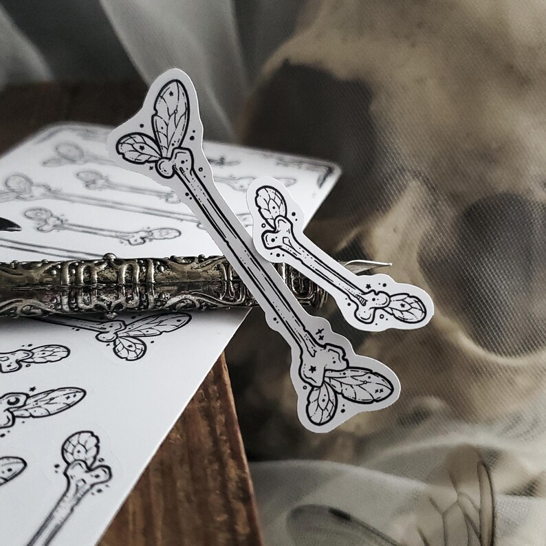 May include: Black and white bone-shaped stickers with leaf-like details. The stickers are on a white surface, with a decorative metal object and a blurred background. The stickers have a whimsical, gothic aesthetic.