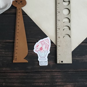 May include: A sticker depicting a pink skull-shaped ice cream sundae on a pedestal. A wooden ruler with a cat head design and a wooden ruler with moon phase cutouts are also present. The background is a dark wood surface.