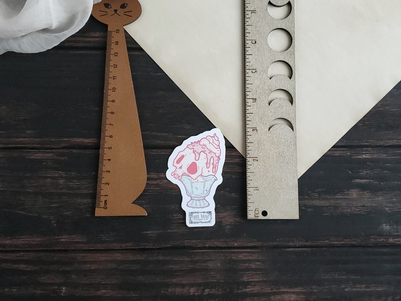 May include: A sticker depicting a pink skull-shaped ice cream sundae on a pedestal. A wooden ruler with a cat head design and a wooden ruler with moon phase cutouts are also present. The background is a dark wood surface.
