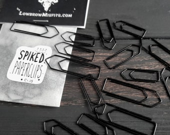 May include: A collection of black spiked paperclips scattered on a dark wooden surface. A small package with the text "SPIKED PAPERCLIPS" and "Cr.15" is visible, along with a card that reads "LOWBROW MISFITS.COM". The paperclips have a unique, angular design.