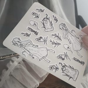 May include: A sheet of black and white stickers featuring a variety of spooky and whimsical designs, including ghosts, flowers, and tombstones.