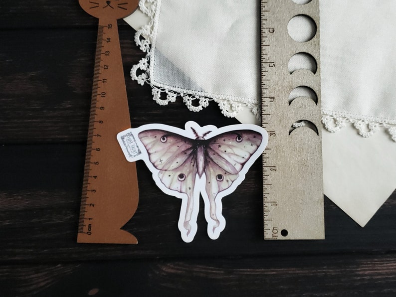 May include: A sticker of a detailed watercolor illustration of a moth with a light brown and purple color scheme. The moth has large, feathery antennae and a wingspan of approximately 5 inches.