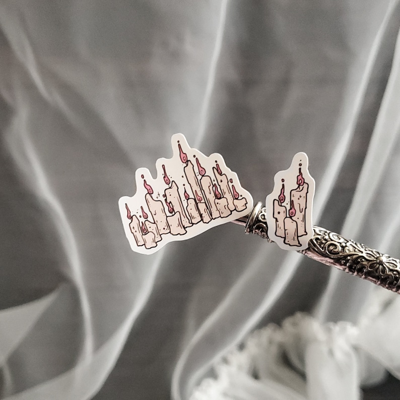May include: Two white sticker sheets with illustrations of candles. The candles are arranged in a row and have pink flames. The stickers are on a silver pen.