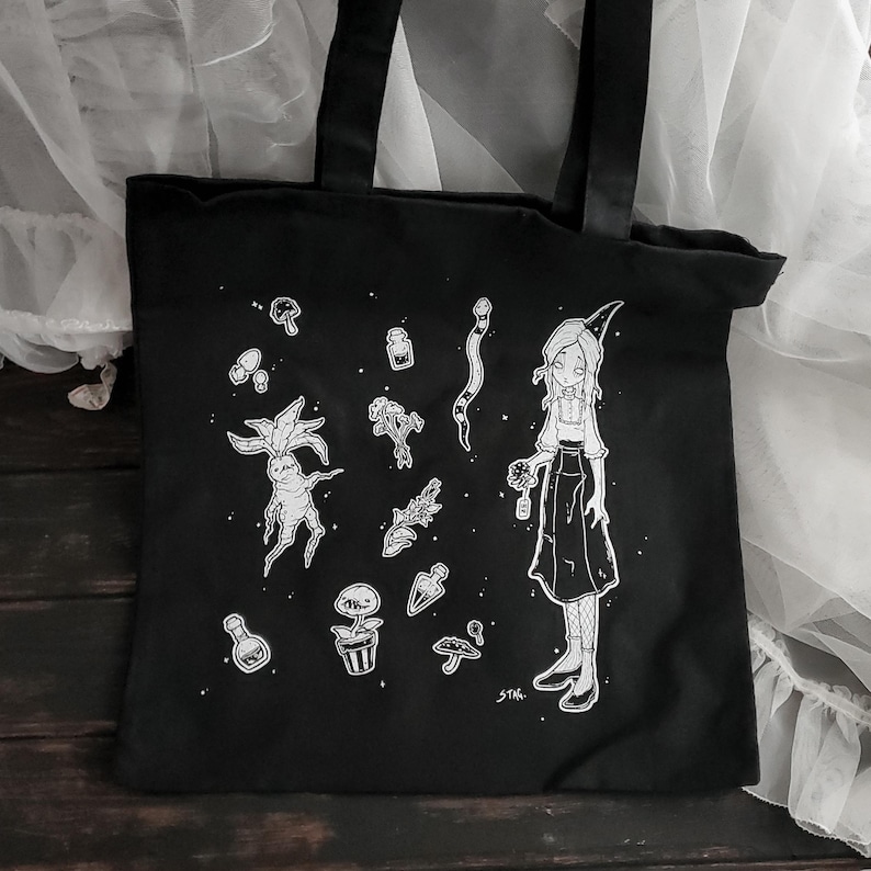 K&ouml;nnte beinhalten: Schwarze Canvas-Tragetasche mit einer wei&szlig;en Illustration einer Hexe, die eine Blume h&auml;lt, umgeben von verschiedenen botanischen Elementen, darunter Pilze, Pflanzen und eine Schlange. Der Text "STAR" befindet sich in der rechten unteren Ecke.