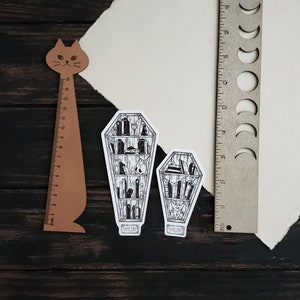 K&ouml;nnte beinhalten: Zwei schwarz-wei&szlig;e Illustrationen von B&uuml;cherregalen in Sargform. Die B&uuml;cherregale sind mit B&uuml;chern und anderen Gegenst&auml;nden gef&uuml;llt. Der Text "Read Until You Die" ist auf der Unterseite jeder Illustration gedruckt.