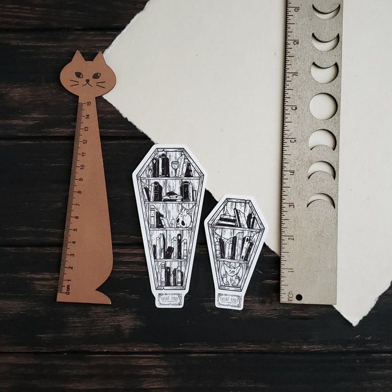 K&ouml;nnte beinhalten: Zwei schwarz-wei&szlig;e Illustrationen von B&uuml;cherregalen in Sargform. Die B&uuml;cherregale sind mit B&uuml;chern und anderen Gegenst&auml;nden gef&uuml;llt. Der Text "Read Until You Die" ist auf der Unterseite jeder Illustration gedruckt.