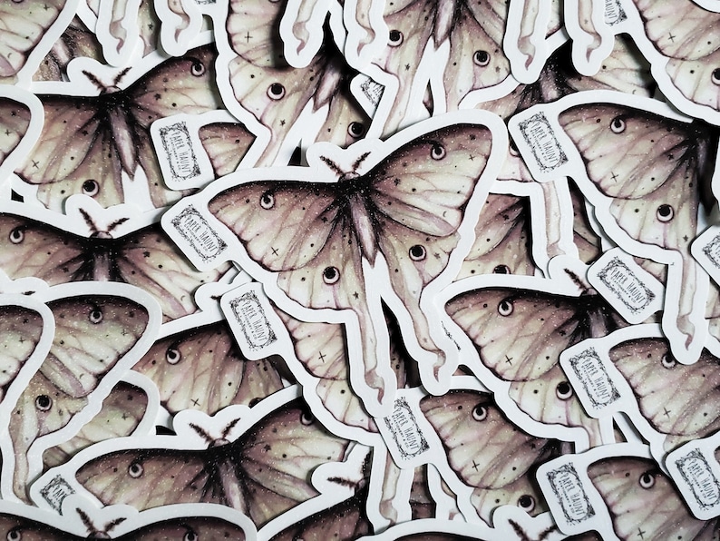 May include: A close-up of a pile of stickers featuring a detailed illustration of a moth with a light brown and beige color scheme. The moth has large, prominent eyes and a delicate, intricate wing pattern. The stickers are printed with the text "Pale Haunt" in a decorative font.