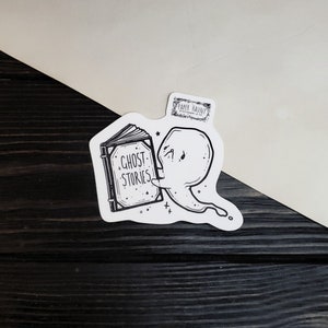 May include: A black and white sticker of a ghost reading a book that says "Ghost Stories".
