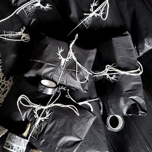 Op de afbeelding: Een stapel zwarte cadeauzakken met wit touw en een sticker van een hert op elke zak. De zakken liggen op een donker houten oppervlak met een decoratieve hand en een rol washi tape.