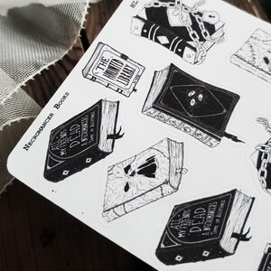 May include: A sheet of stickers featuring black and white illustrations of books with spooky titles like "All My Friends Are Dead" and "The Haunted Library". The stickers are designed in a gothic style with intricate details and are perfect for adding a touch of macabre to your planner, laptop, or water bottle.