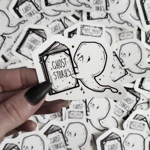 May include: A black and white sticker featuring a ghost reading a book with the text "Ghost Stories" on the cover. The sticker is on a white background and has a small logo that says "Paper Haunt".