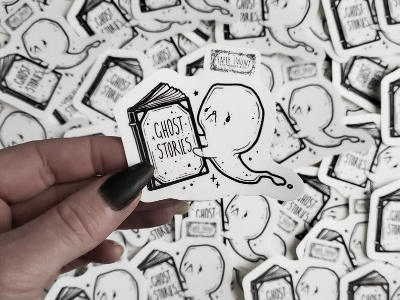 May include: A black and white sticker featuring a ghost reading a book with the text "Ghost Stories" on the cover. The sticker is on a white background and has a small logo that says "Paper Haunt".