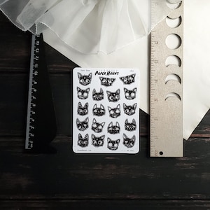 May include: A sheet of black and white cat face stickers with the text "Paper Haunt" in a white banner. The stickers are arranged in a grid pattern and feature various expressions.