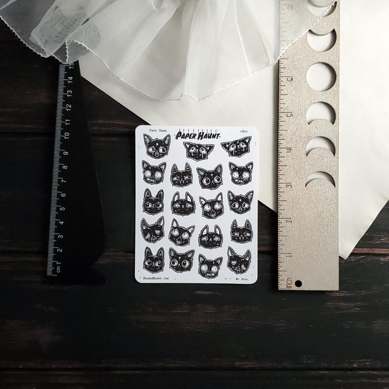 May include: A sheet of black and white cat face stickers with the text "Paper Haunt" in a white banner. The stickers are arranged in a grid pattern and feature various expressions.