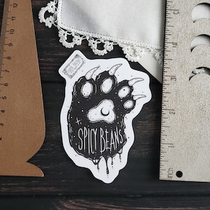 Puede incluir: Una pegatina en blanco y negro con una huella de pata de oso estilizada y el texto "SPICY BEANS". La pegatina mide aproximadamente 7.6 cm de largo y est&aacute; colocada sobre una superficie de madera oscura. Tambi&eacute;n se ven una regla marr&oacute;n y una regla plateada.