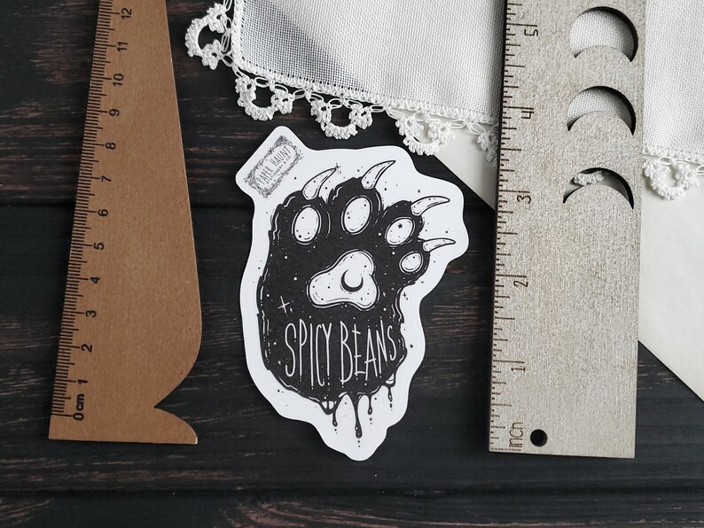 Puede incluir: Una pegatina en blanco y negro con una huella de pata de oso estilizada y el texto "SPICY BEANS". La pegatina mide aproximadamente 7.6 cm de largo y est&aacute; colocada sobre una superficie de madera oscura. Tambi&eacute;n se ven una regla marr&oacute;n y una regla plateada.
