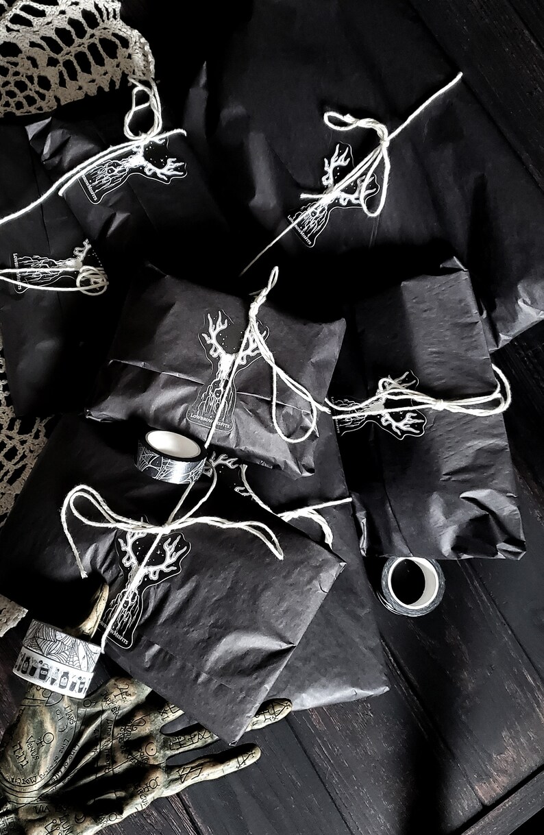 May include: A pile of black gift bags with white string and a white deer sticker on each bag. The bags are on a dark wooden surface with a decorative hand and a roll of tape.