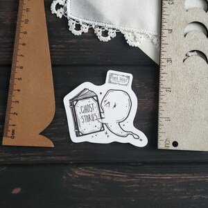 May include: A black and white sticker of a ghost reading a book that says "Ghost Stories". The sticker is on a wooden surface.