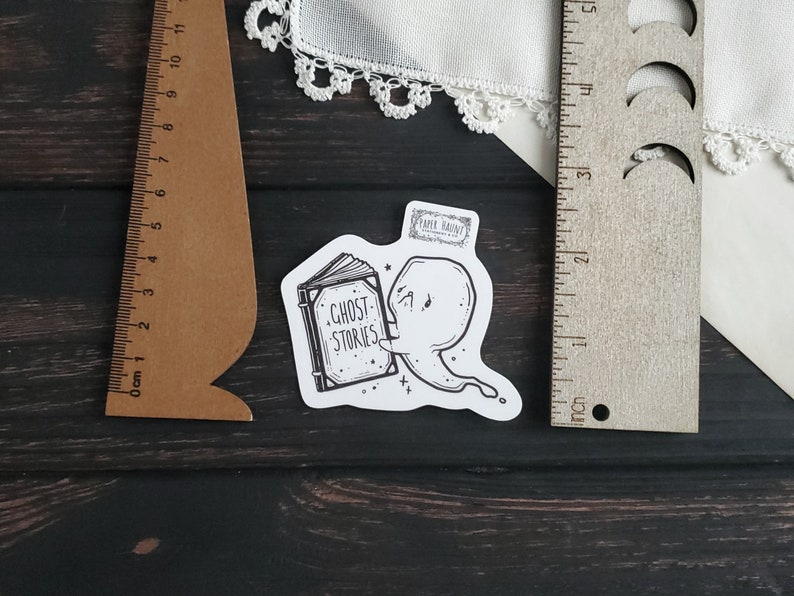 May include: A black and white sticker of a ghost reading a book that says "Ghost Stories". The sticker is on a wooden surface.