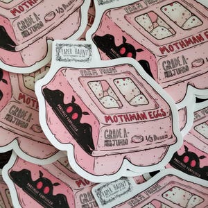 Puede incluir: Pegatina rosa que representa una huevera con la inscripción "Mothman Eggs". La huevera lleva las etiquetas "Grade A Jumbo" y "Farm Fresh". Una silueta negra de criatura parecida a una polilla está en el lateral. La pegatina es de "Paper Haunt Stationery & Co."