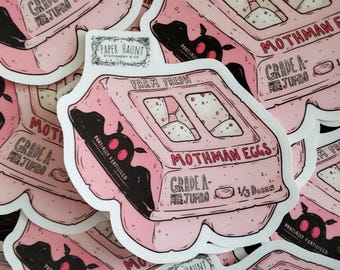 May include: Pink sticker featuring a cartoon egg carton labeled "Mothman Eggs." The carton reads "Grade A Jumbo" and "Farm Fresh." A black silhouette of a moth-like creature is on the side. The sticker is from "Paper Haunt Stationery & Co."