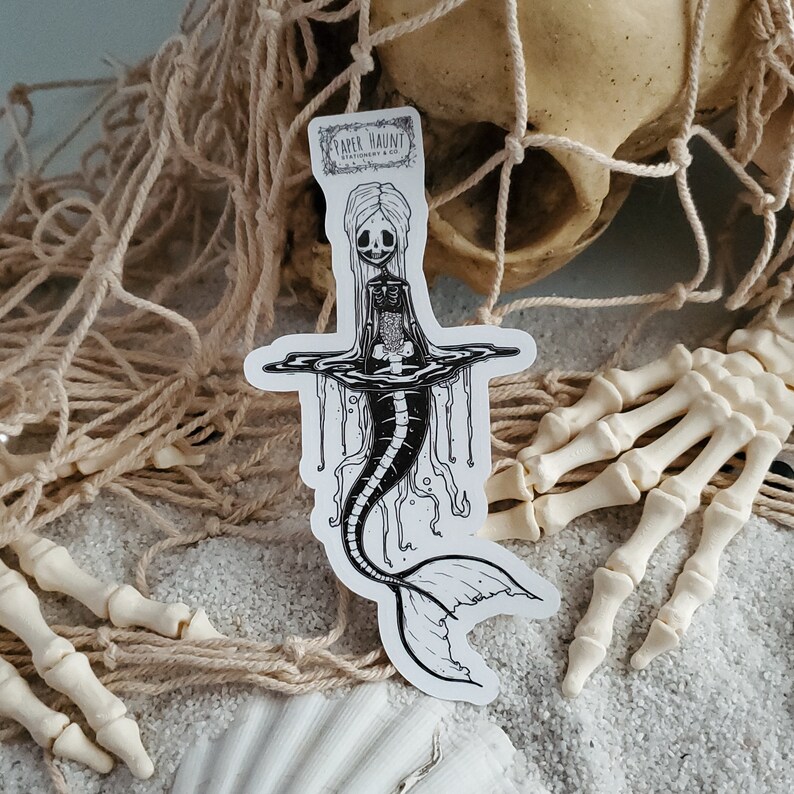 Spooky cute goth Jelly bones mermaid skeleton emerging from water, a sticker Featuring a Mermaid with Translucent skin with visible skeleton and organs pictured on sand with a skull and skeleton arms reaching from a fishing net.