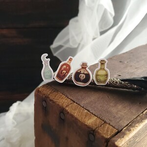 May include: Four small glass bottles with cork stoppers and colorful liquids. The bottles are arranged in a row on a wooden surface. The bottles are decorated with a crescent moon, a skull and crossbones, and other designs.