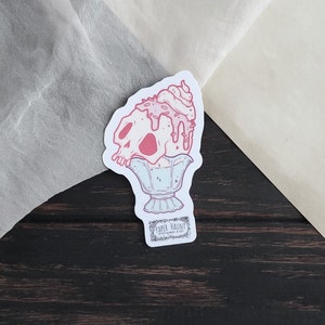 May include: A sticker depicting a pink skull sundae with dripping ice cream and whipped cream. The sundae is in a decorative glass, with the text "PAPER HAUNT STATIONERY & CO." at the bottom. The sticker has a white border.