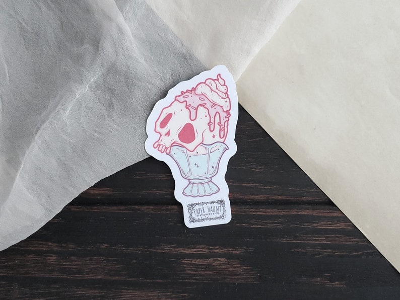 May include: A sticker depicting a pink skull sundae with dripping ice cream and whipped cream. The sundae is in a decorative glass, with the text "PAPER HAUNT STATIONERY & CO." at the bottom. The sticker has a white border.