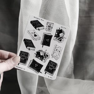 May include: A sheet of black and white stickers featuring illustrations of books with titles like "Necromancer Books", "The Haunted Library", "110 Rules for Conquering Dead", and "Mysteries of the Dead".