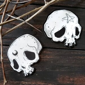 Op de afbeelding: Twee zwart-witte schedelpatches met verschillende ontwerpen. De ene schedel heeft een halve maan en de andere een pentagram.