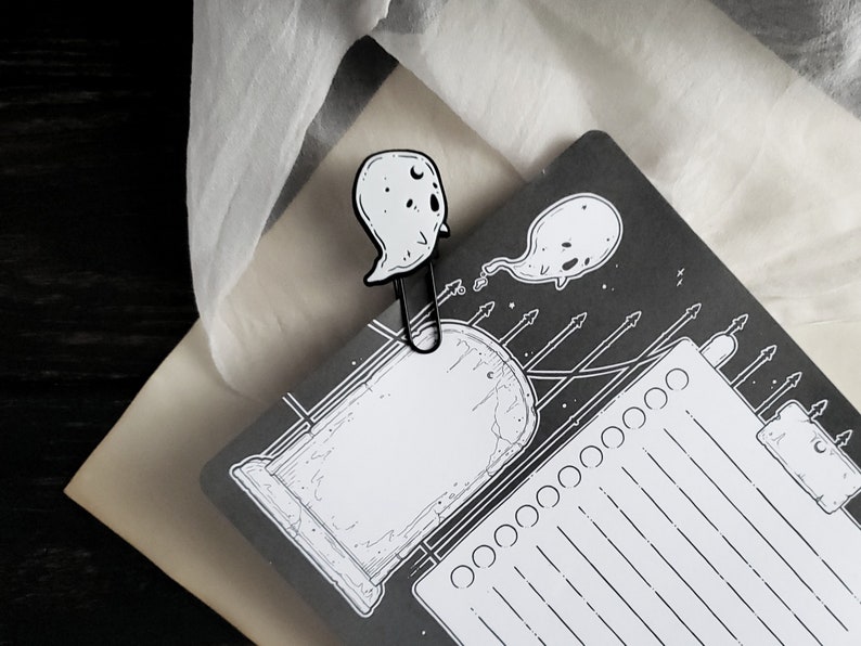 May include: A black and white illustrated ghost bookmark with a black metal clip. The bookmark features a ghost with a crescent moon on its head and a smaller ghost with a tail.
