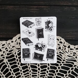 May include: A sheet of black and white stickers featuring illustrations of books with spooky titles like "The Haunted Library", "All The Dead We Remember", and "110 Rules of Summoning the Dead".