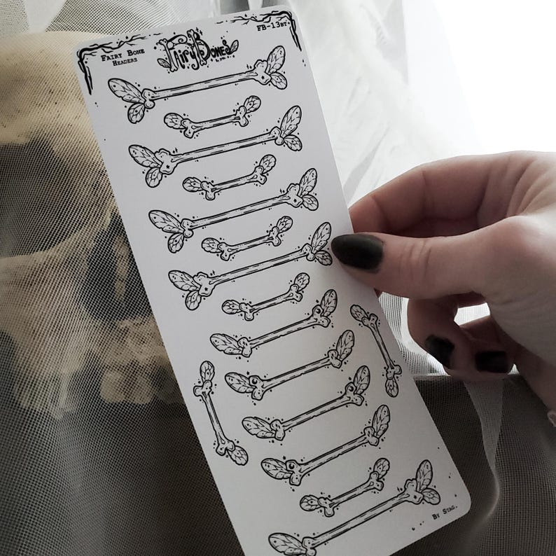May include: A white sticker sheet featuring black illustrations of stylized bones with leaf-like accents. The sheet is labeled "Fairy Bone Headers" and includes the text "FB-13wt". The sticker sheet is held by a hand with dark nail polish.