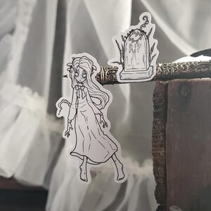 May include: Black and white line art sticker set featuring a spooky girl in a dress and a tombstone with the inscription "RIP".