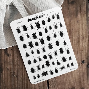 May include: A white sticker sheet featuring numerous black beetle illustrations of varying sizes. The sheet includes the text "Paper Haunt" and "Beetles". The sticker sheet is on a wooden surface.