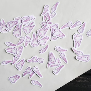 May include: A collection of pink and purple tooth stickers. The stickers are scattered on a white surface. The stickers are in various shapes and sizes.