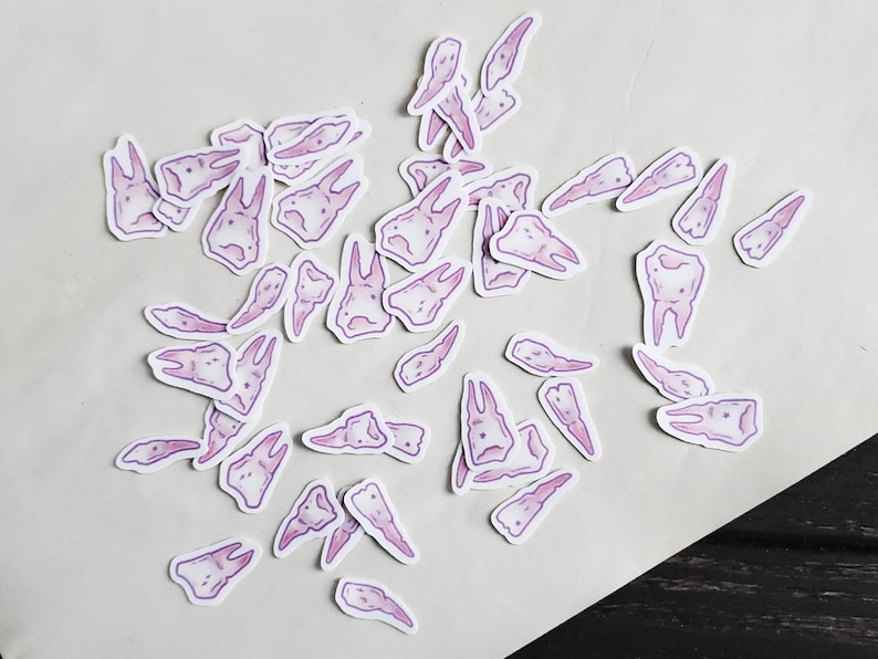 May include: A collection of pink and purple tooth stickers. The stickers are scattered on a white surface. The stickers are in various shapes and sizes.