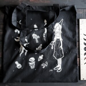 K&ouml;nnte beinhalten: Schwarze Tragetasche mit einem wei&szlig;en Design, das eine Hexe zeigt, die eine Blume h&auml;lt, eine Schlange und verschiedene andere botanische Illustrationen. Die Tasche hat zwei Henkel und der Text "STAG." ist in der rechten unteren Ecke gedruckt.