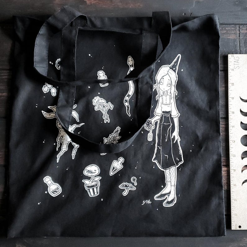 K&ouml;nnte beinhalten: Schwarze Tragetasche mit einem wei&szlig;en Design, das eine Hexe zeigt, die eine Blume h&auml;lt, eine Schlange und verschiedene andere botanische Illustrationen. Die Tasche hat zwei Henkel und der Text "STAG." ist in der rechten unteren Ecke gedruckt.