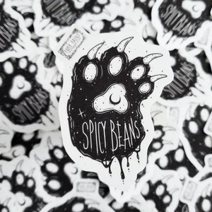 Puede incluir: Pegatinas en blanco y negro con un dise&ntilde;o de huella de oso y la frase "SPICY BEANS". El dise&ntilde;o incluye una luna creciente y estrellas. Las pegatinas est&aacute;n dispersas sobre una superficie blanca.