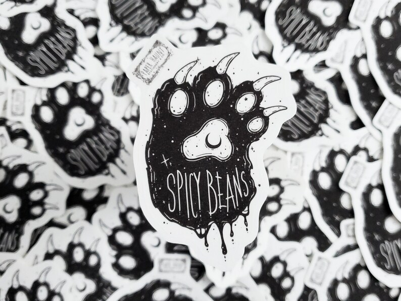 Puede incluir: Pegatinas en blanco y negro con un dise&ntilde;o de huella de oso y la frase "SPICY BEANS". El dise&ntilde;o incluye una luna creciente y estrellas. Las pegatinas est&aacute;n dispersas sobre una superficie blanca.