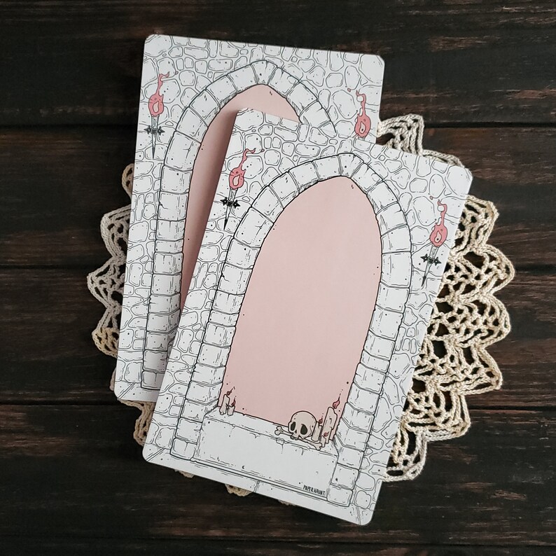 Pode incluir: Dois cart&otilde;es imprim&iacute;veis em branco com um arco de pedra e um design de caveira. Os cart&otilde;es s&atilde;o brancos com contornos pretos e um fundo rosa dentro do arco. O texto do cart&atilde;o diz "Paper & Ink Co."