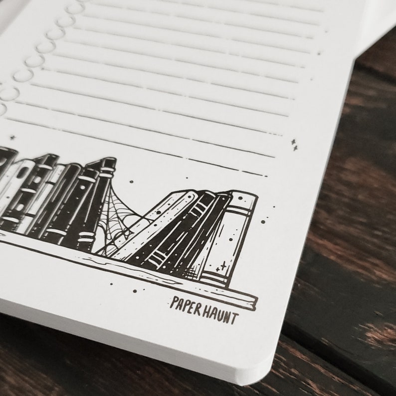 K&ouml;nnte beinhalten: Ein wei&szlig;es Notizbuch mit einer schwarz-wei&szlig;en Illustration eines B&uuml;cherregals mit B&uuml;chern und einem Spinnennetz. Der Text "PAPER HAUNT" ist unten auf dem Notizbuch gedruckt.
