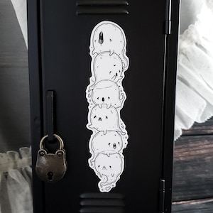 May include: A white sticker featuring a stack of five cartoon ghosts with various expressions. The sticker is placed on a black locker door with a vintage padlock. The ghosts have simple, rounded shapes and small, dark eyes.