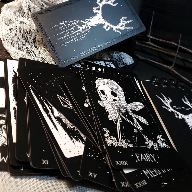 May include: A deck of black tarot cards with white illustrations. The cards feature a variety of images, including a fairy, a skull, and a deer. The cards are numbered with Roman numerals and have the word "Fairy" written on one of the cards.