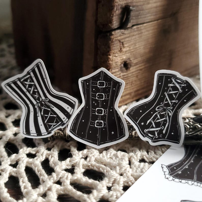 May include: Three black and white corset stickers. The corsets are in different styles, one with stripes, one with buckles, and one with lace. The stickers are on a white surface.