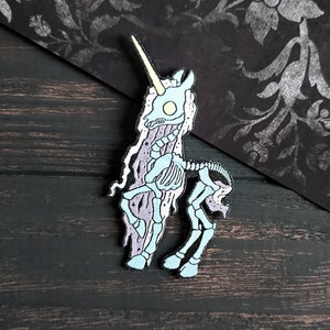 Op de afbeelding: Een blauwe en witte emaille pin van een skeletachtig eenhoorn met een gele hoorn. Het eenhoorn staat op een donker houten oppervlak.