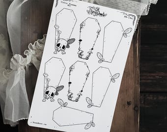 May include: A white sticker sheet featuring coffin-shaped designs with skull and crossbones, mushrooms, and leaf accents. The text "Coffin Planner Box" and "Fairy Bones" are visible. The sheet is on a wooden surface.
